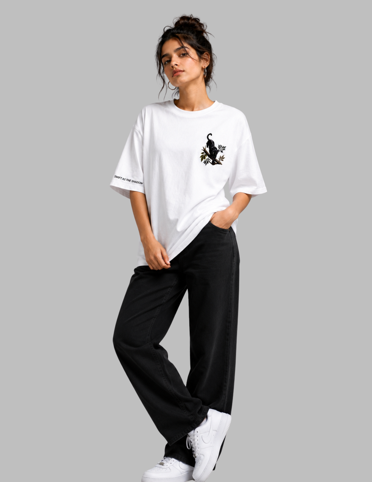 A woman modeling a white oversized t-shirt with a black cat graphic on the chest and black wide-leg pants, standing against a solid gray background.