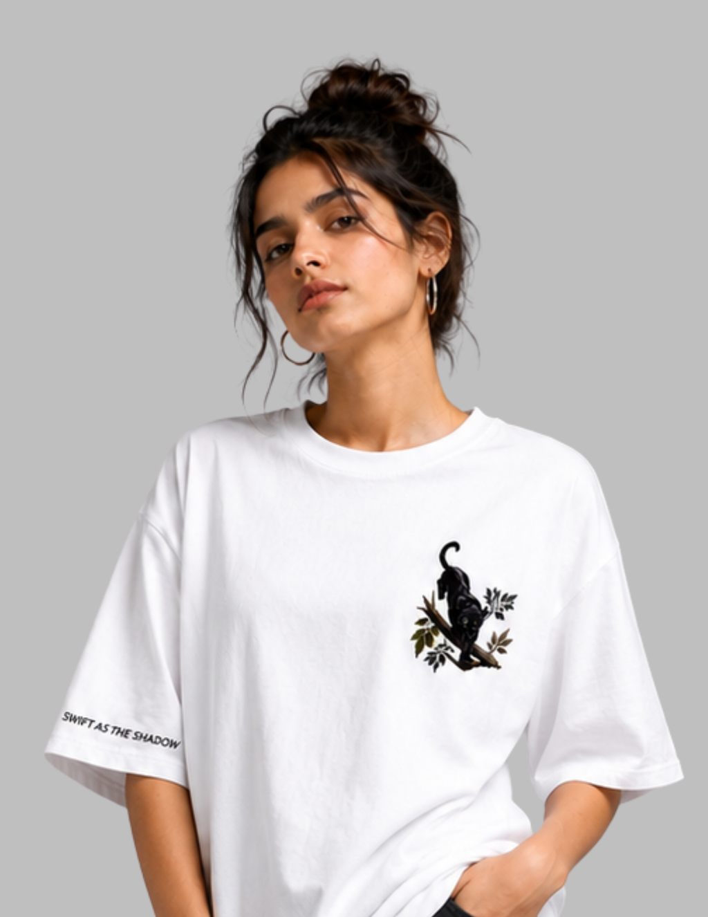 A woman wearing an oversized white t-shirt with a black cat graphic on the left chest and 'SHYFT AS THE SHADOW' text on the sleeve, set against a solid gray background.