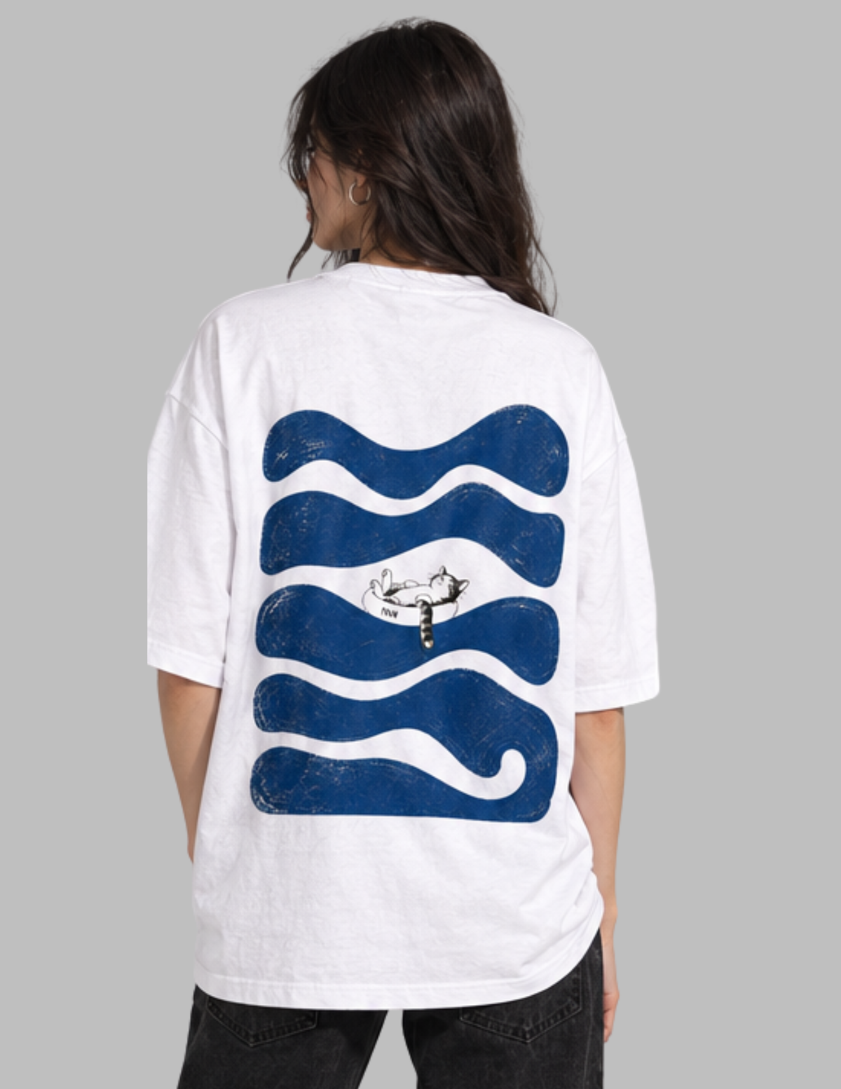 A person shown from the back wearing an oversized white T-shirt with a large blue wavy graphic and a small central illustration, paired with dark pants, against a plain light gray background.