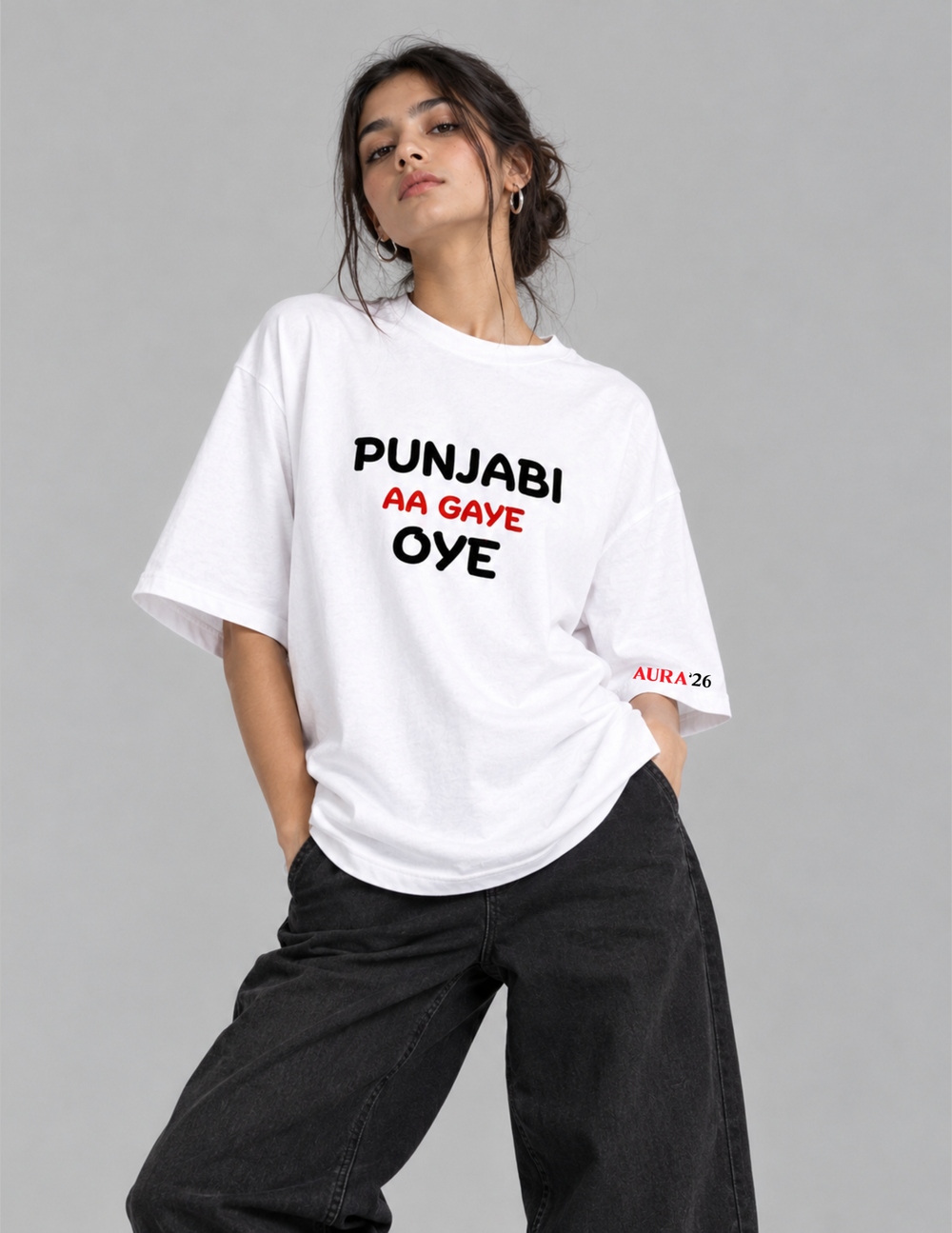 A woman modeling an oversized white graphic T-shirt with bold black and red text reading 'PUNJABI AA GAVE OYE' on the front, paired with loose black pants against a plain gray studio background.