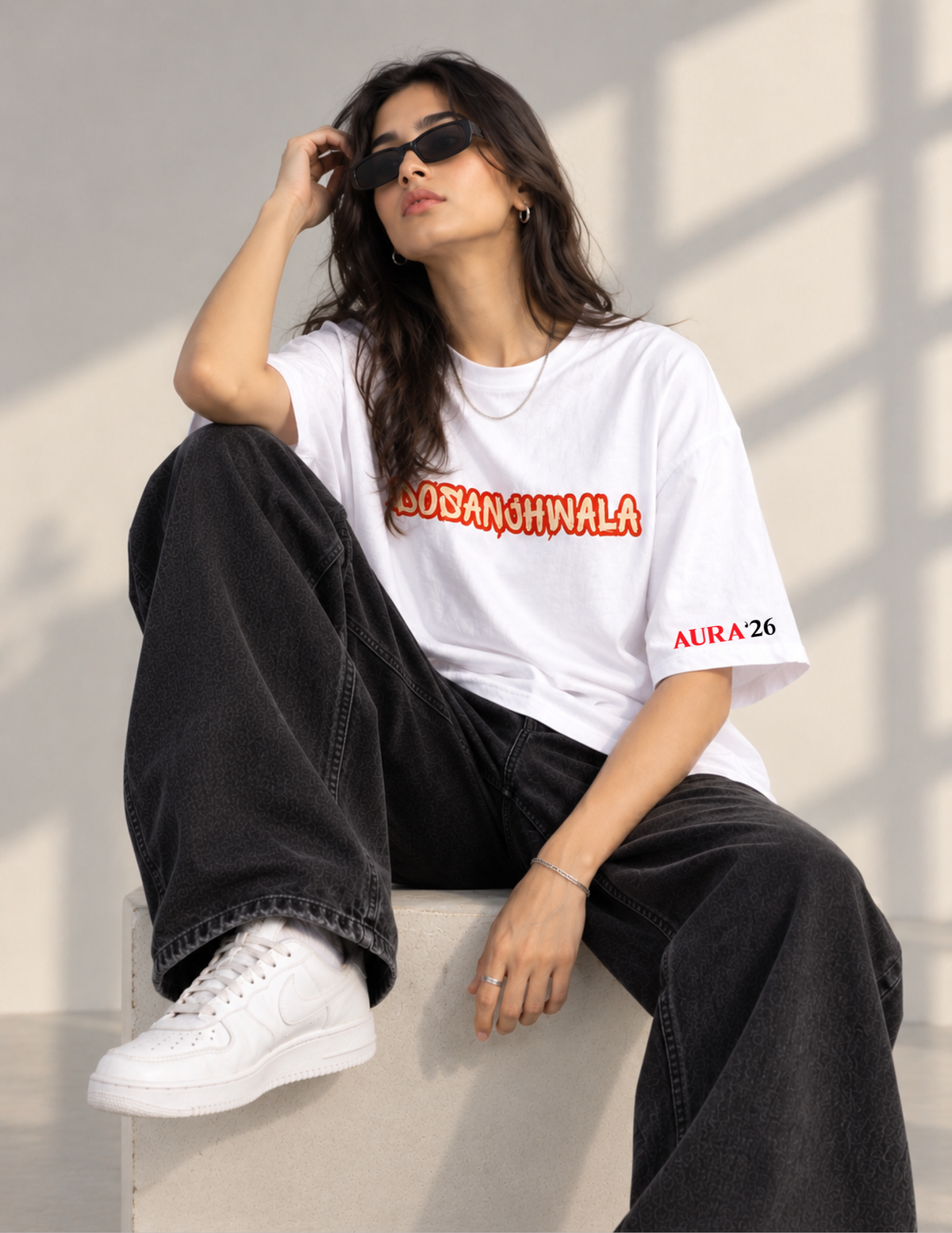 A woman wearing a white oversized graphic T-shirt with red and orange text reading 'DOSAN CHIWALA' across the chest and 'AURA 26' on the sleeve, paired with black wide-leg jeans and white sneakers, seated against a sunlit wall.