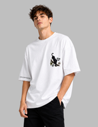 A man wearing an oversized white t-shirt with a black cat graphic on the left chest and text on the sleeve, paired with black pants, against a solid gray background.