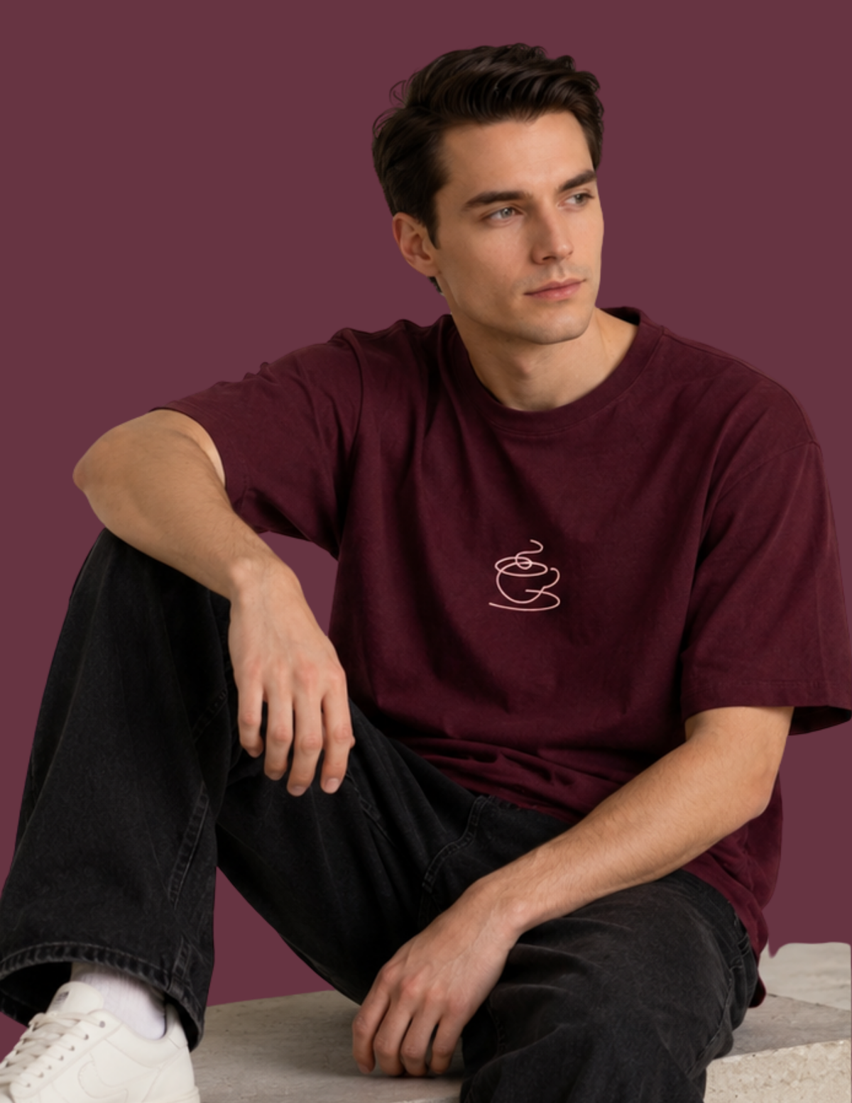 A man wearing a burgundy t-shirt with a small white coffee cup graphic on the chest, paired with black jeans and white sneakers, sitting against a solid burgundy background.