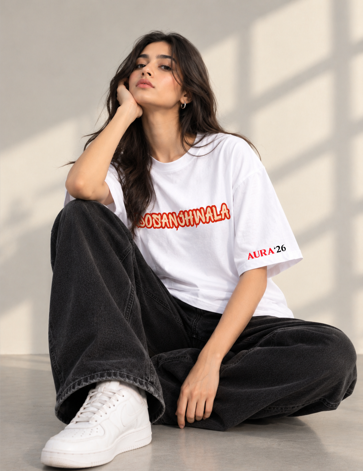 A woman sits cross-legged on a concrete floor wearing a white t-shirt with red text reading 'DOBANSHINALA' and 'ALIRA 26' on the sleeve, paired with black wide-leg jeans and white sneakers.