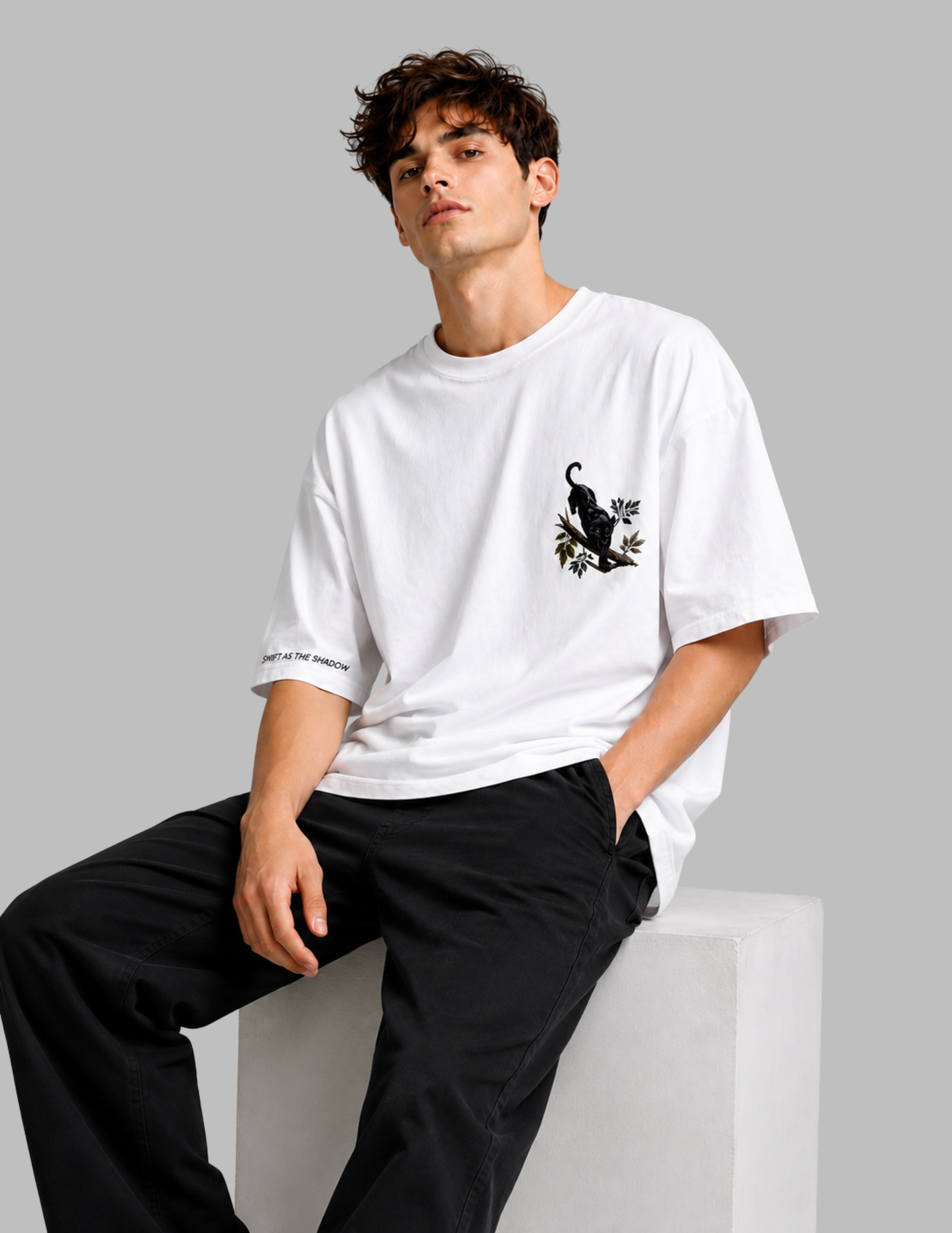 A man wearing a white short-sleeve t-shirt with a black cat graphic on the left chest, paired with black pants, sitting on a white block against a solid gray background.