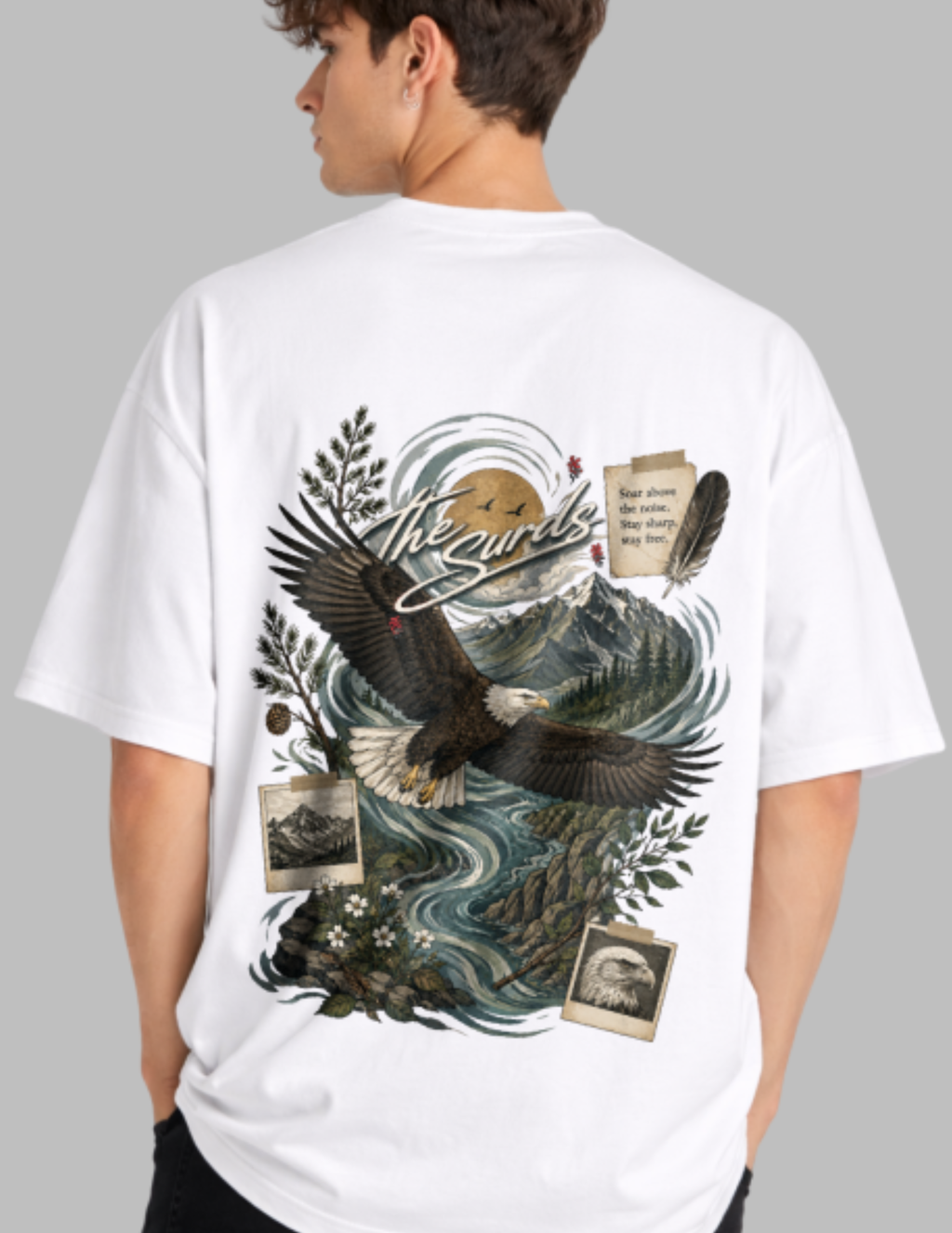 A man wearing a white oversized T-shirt, shown from the back. The shirt features a large, detailed graphic print of an eagle flying over a mountain river landscape with vintage-style photo frames and text.