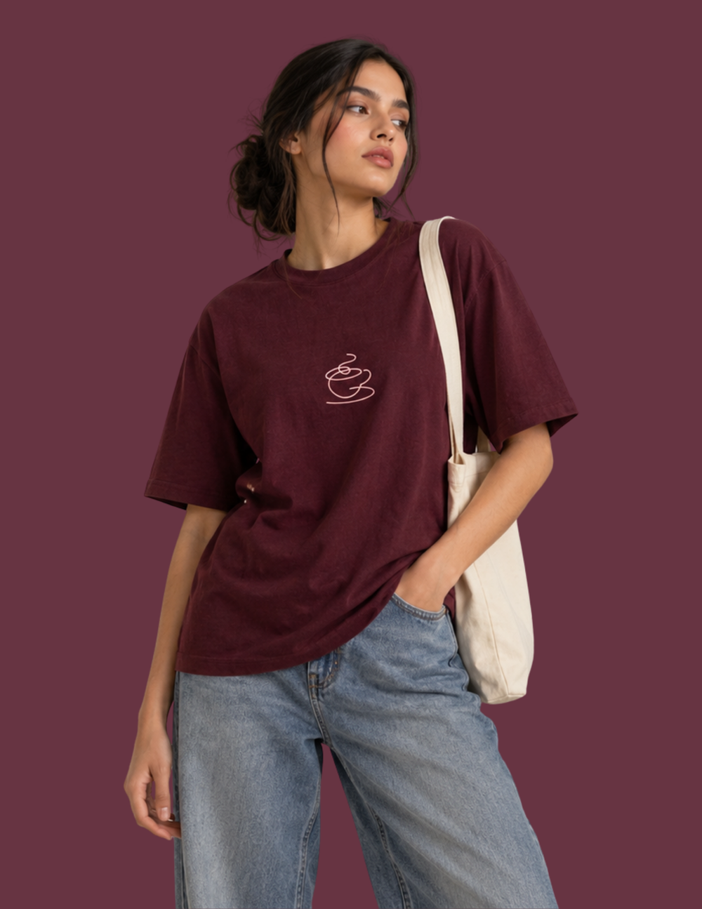 A woman wearing a maroon short-sleeve t-shirt with a small white coffee cup graphic on the chest, paired with light blue jeans and a cream canvas shoulder bag, set against a solid maroon background.