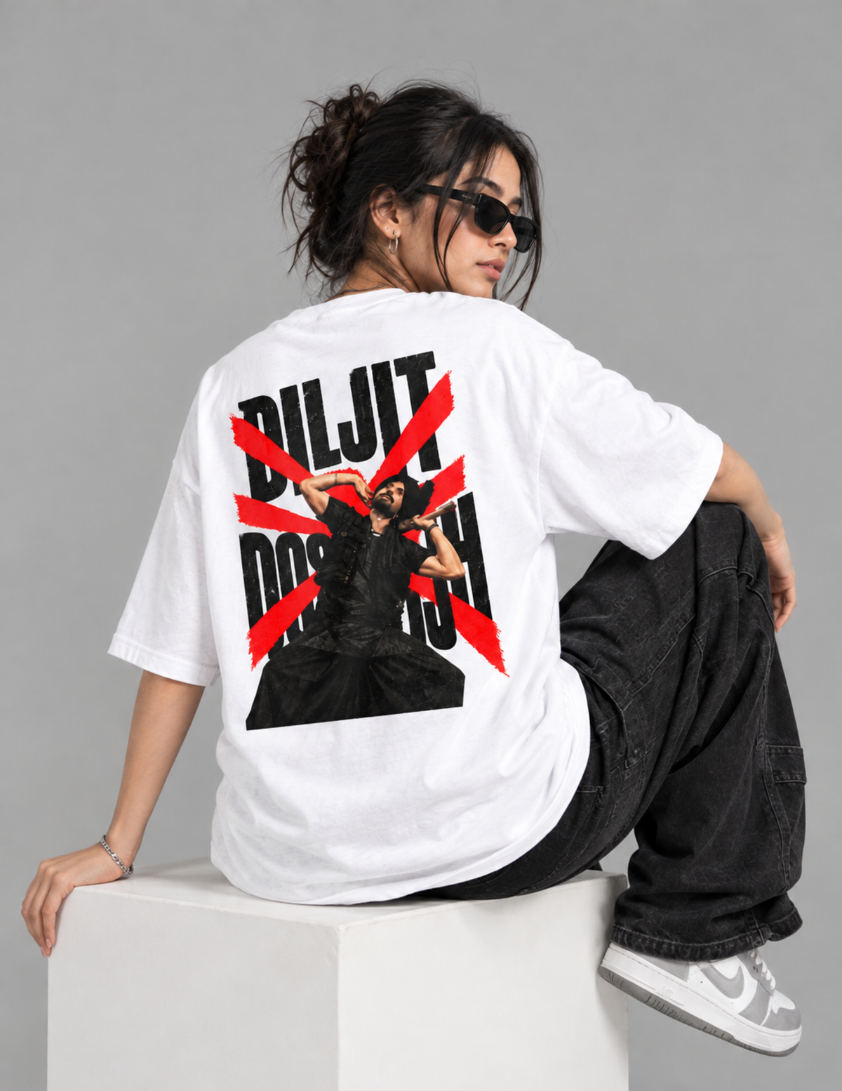 A model wearing a white oversized t-shirt with a large graphic print on the back, sitting on a white block against a gray studio background.