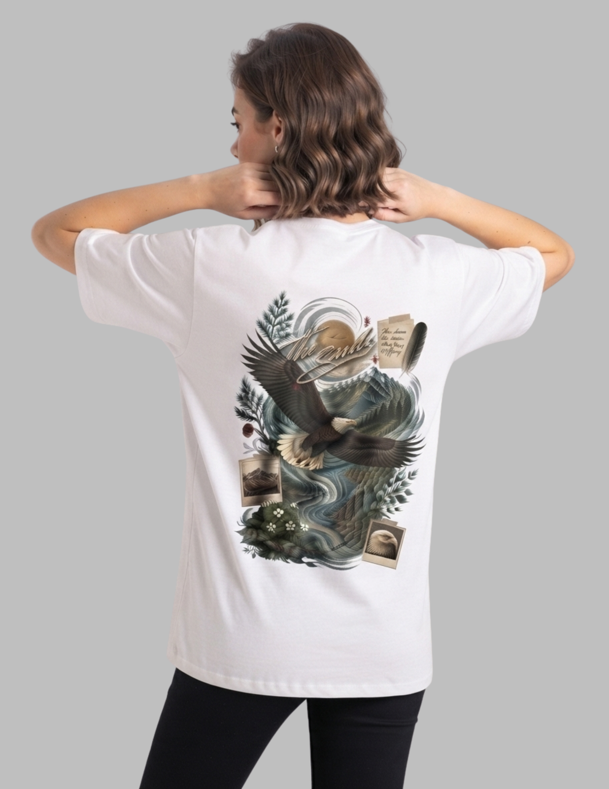 A woman shown from the back wearing a white short-sleeve T-shirt with a large detailed graphic print on the back featuring an eagle, waves, and parchment, paired with black pants against a solid gray background.