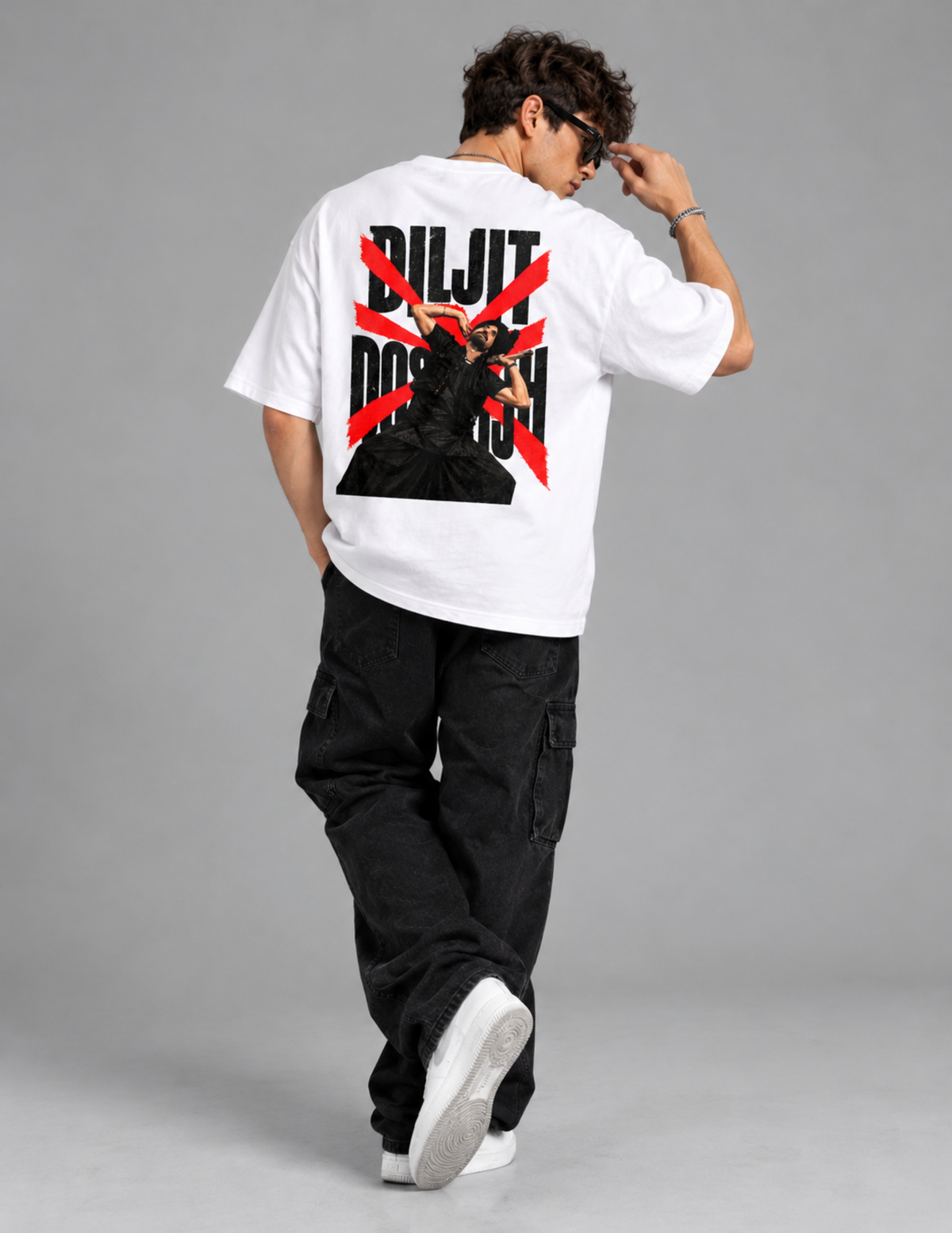 A man wearing a white oversized t-shirt with a large black and red graphic print on the back, paired with black cargo pants and white sneakers, photographed from behind against a solid gray studio background.