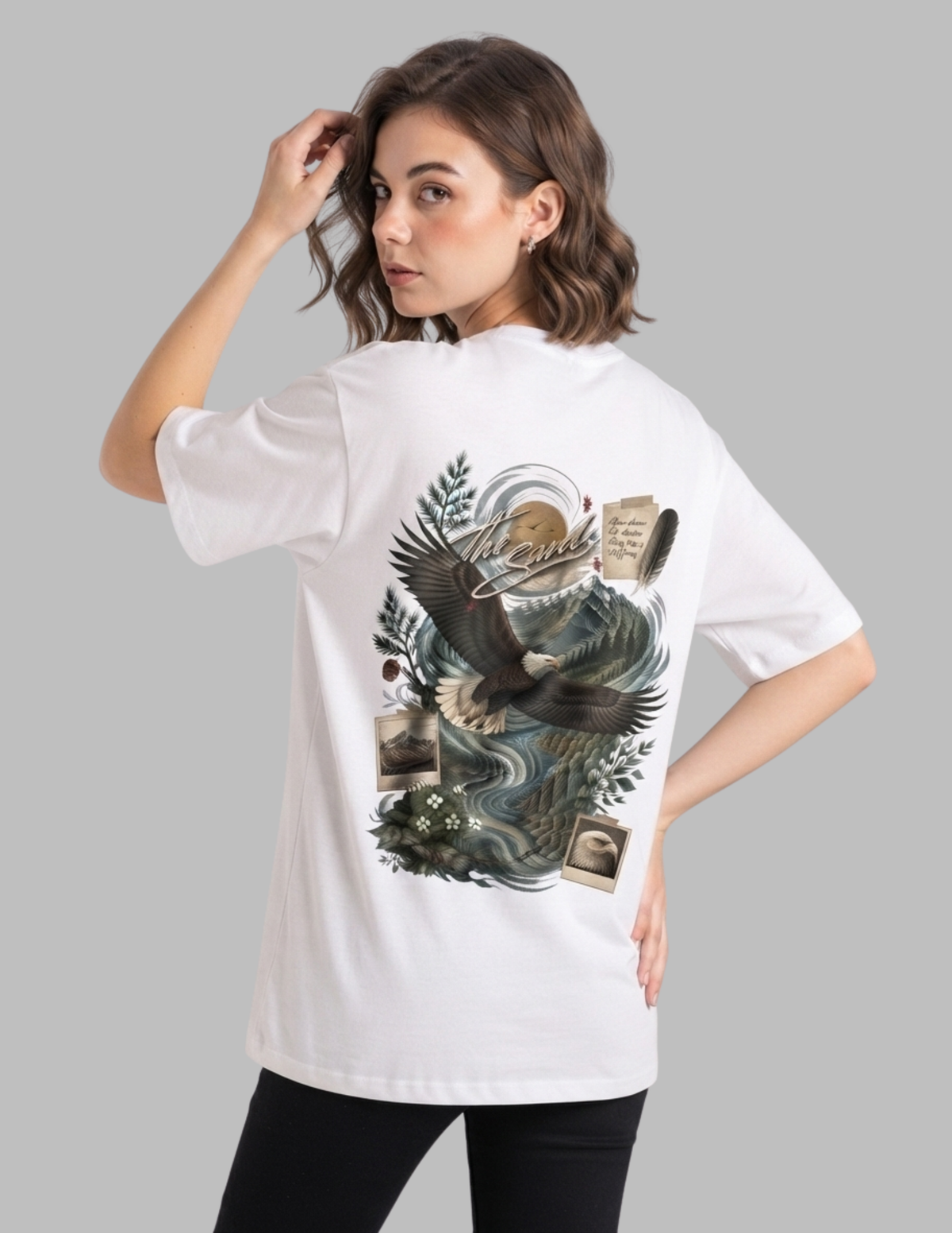 A woman modeling a white oversized T-shirt with a large graphic print on the back, featuring an eagle, waves, and vintage postcard elements, against a solid gray background.