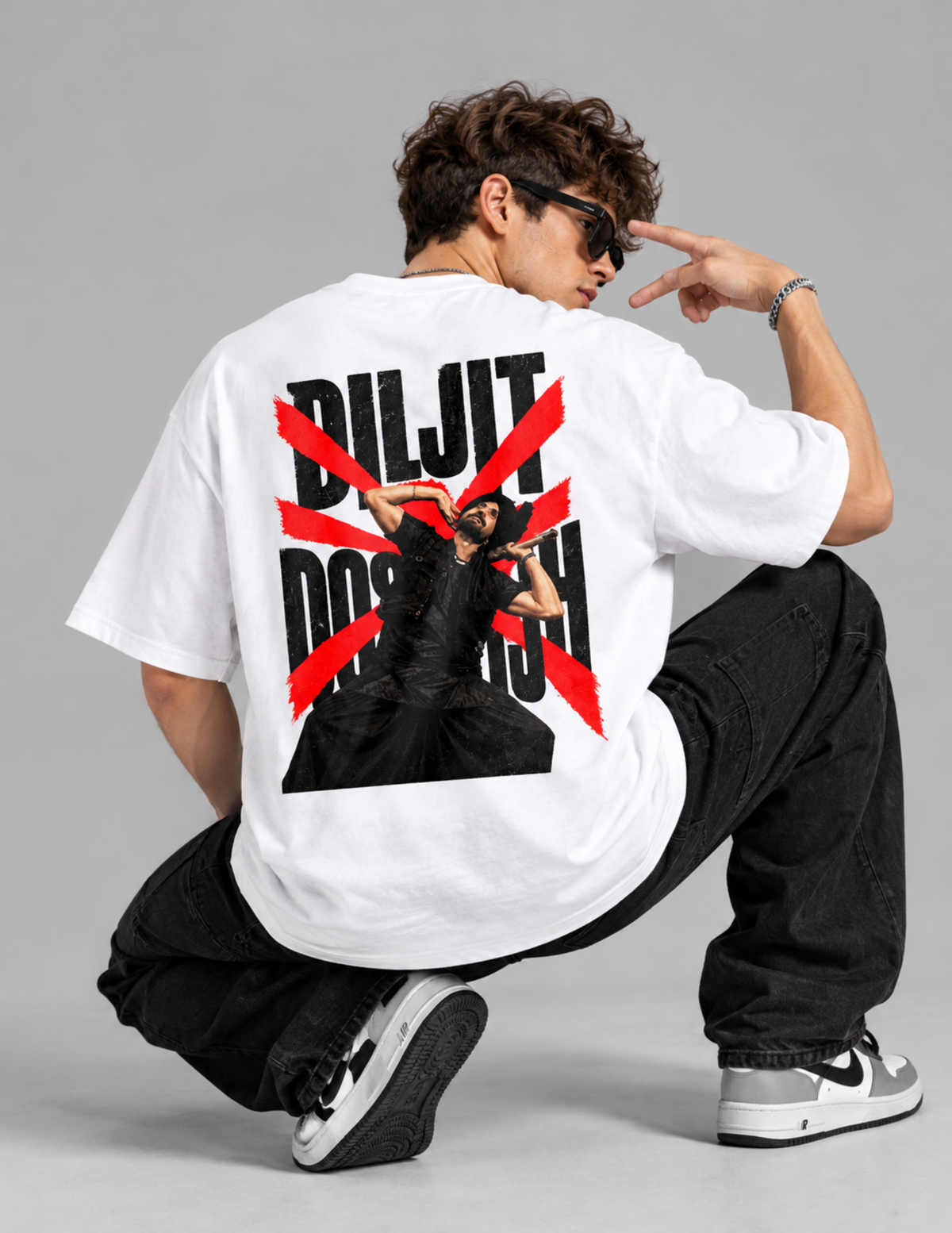 A model wearing a white oversized t-shirt with a large graphic print on the back, crouching in a studio setting. The graphic features the text 'DILJI DOSTOH' in black with a red diagonal X overlay and a black figure in the center.