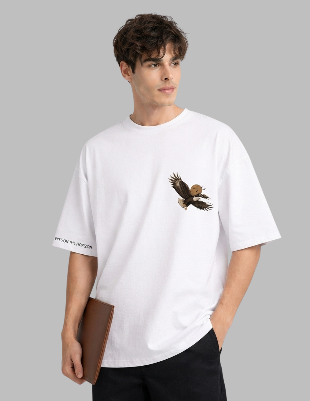 A man wearing a white oversized crewneck t-shirt with a graphic of a flying bald eagle on the left chest and black text on the right sleeve, set against a solid gray background.