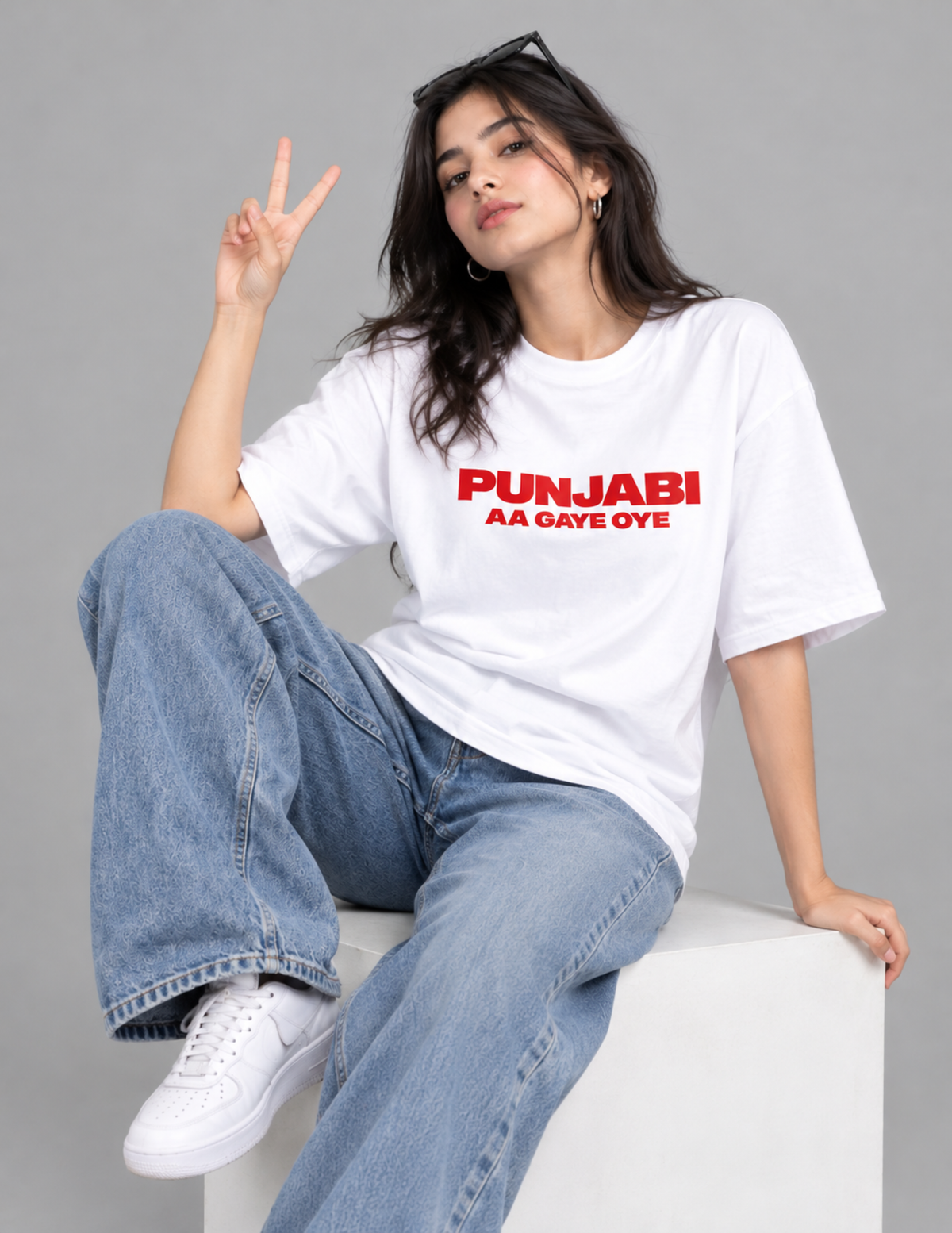 A woman sitting on a white block wearing a white t-shirt with red text that reads 'PUNJABI AA GAYE OYE', paired with wide-leg blue jeans and white sneakers, posing against a gray studio background.