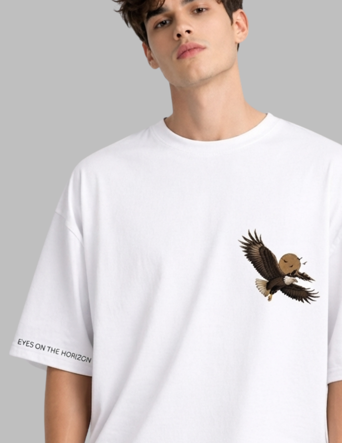 A man wearing a white oversized t-shirt with a brown and black eagle graphic on the left chest and 'EYES ON THE HORIZON' text on the right sleeve, set against a solid gray background.