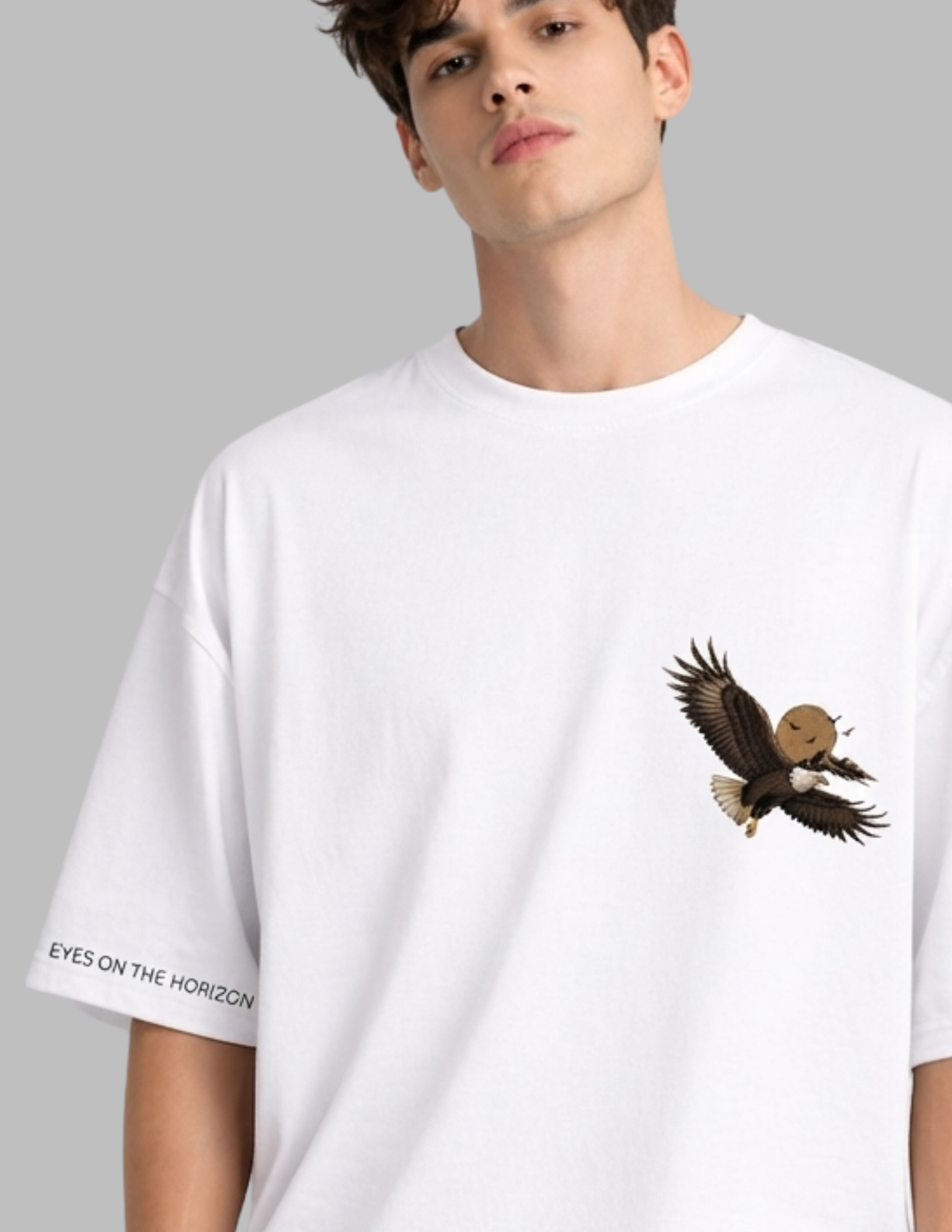 A man wearing a white oversized t-shirt with a brown and black eagle graphic on the left chest and 'EYES ON THE HORIZON' text on the right sleeve, set against a solid gray background.