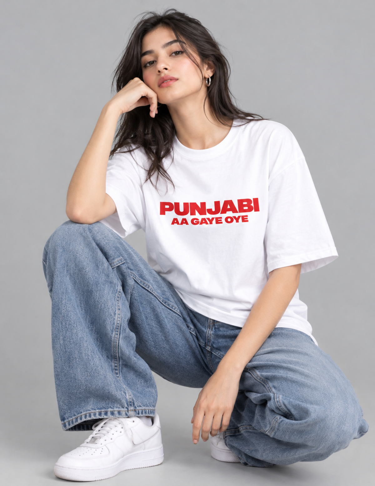 A woman crouching in a studio setting, wearing a white oversized graphic T-shirt with red text reading 'PUNJABI AA GAYE OYE', light blue wide-leg jeans, and white sneakers against a plain gray background.