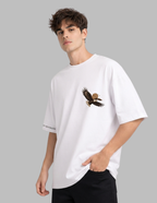 A man wearing a white oversized crewneck t-shirt with a brown and black eagle graphic on the left chest, paired with black pants, set against a solid gray background.