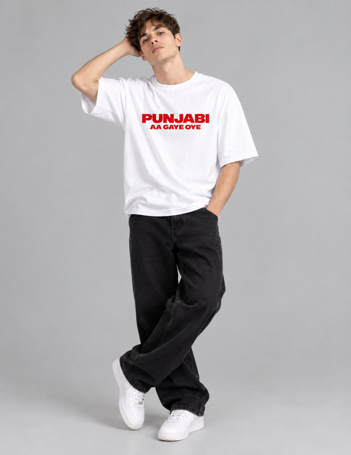 A man modeling a white short-sleeve t-shirt with red text that reads 'PUNJABI AA GATE OYE' on the chest, paired with black jeans and white sneakers, against a solid gray studio background.