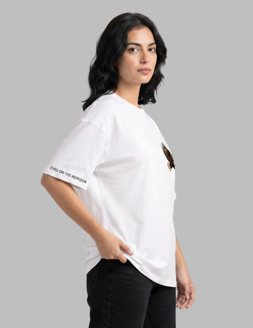 A woman wearing a white oversized t-shirt with a small brown eagle graphic on the left chest and 'EYES ON THE MORIOM' text on the right sleeve, paired with black jeans, set against a solid gray background.