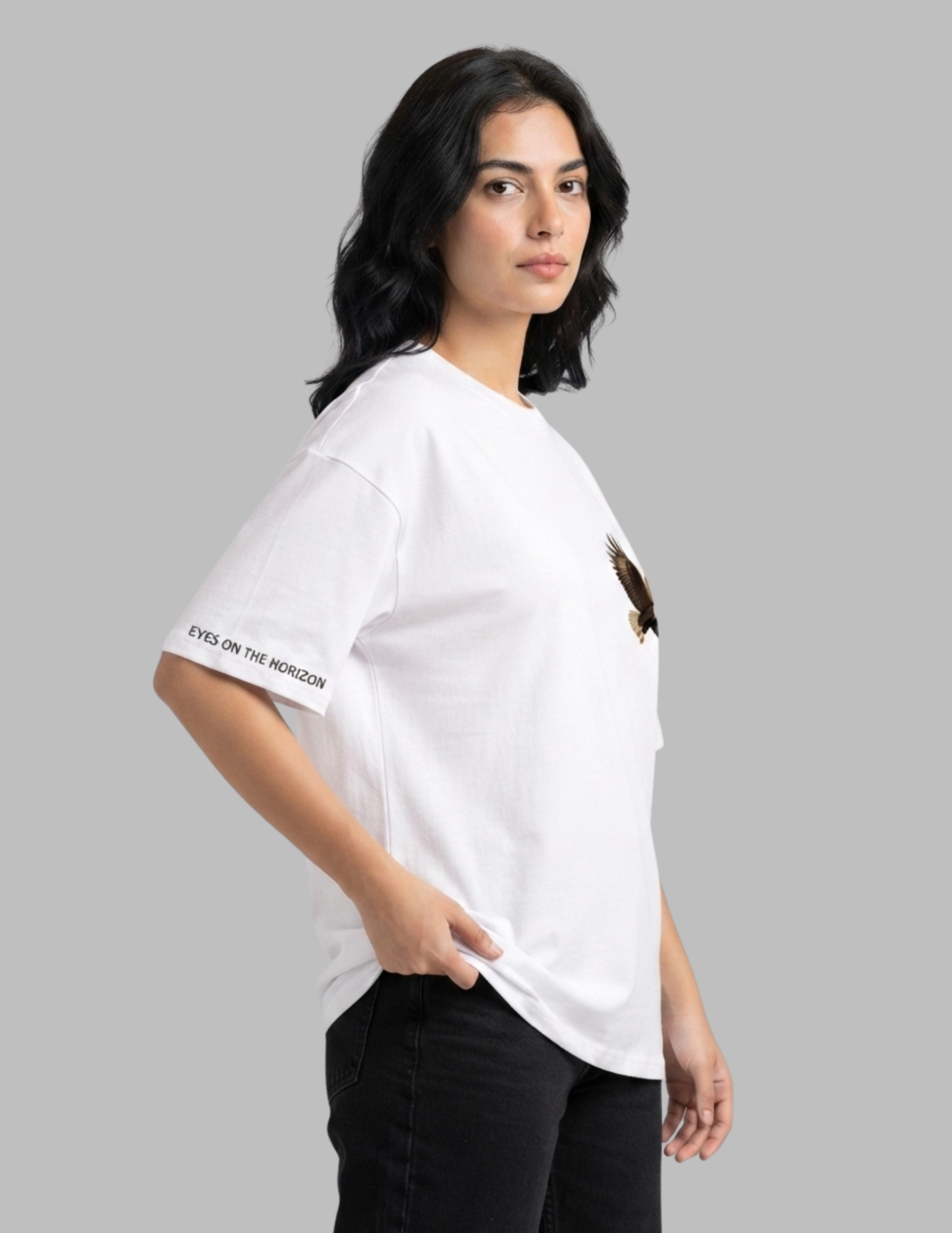 A woman wearing a white oversized t-shirt with a small brown eagle graphic on the left chest and 'EYES ON THE MORIOM' text on the right sleeve, paired with black jeans, set against a solid gray background.