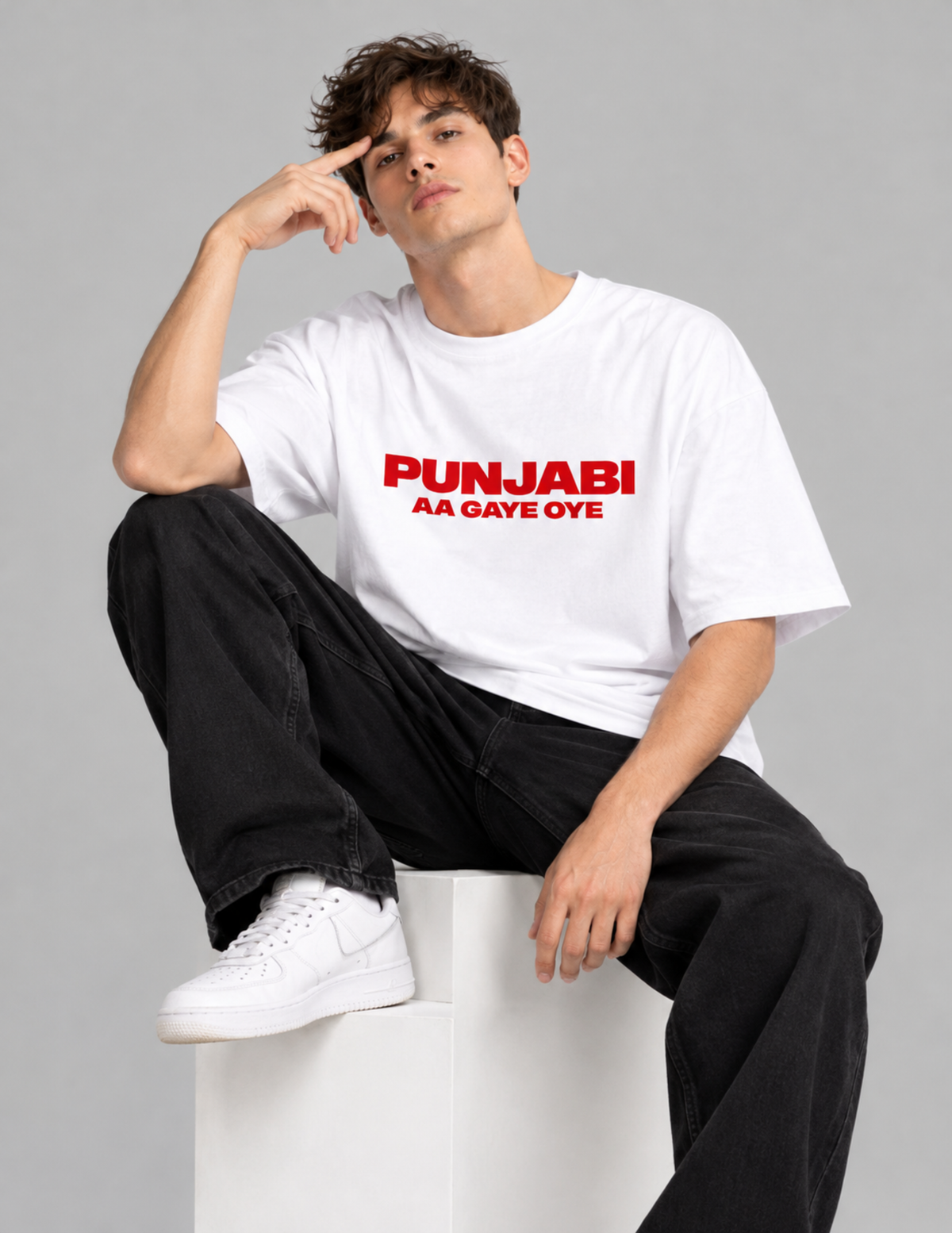 A man wearing a white t-shirt with red text that reads 'PUNJABI AA GAVE OYE' sits on a white block against a gray studio background.