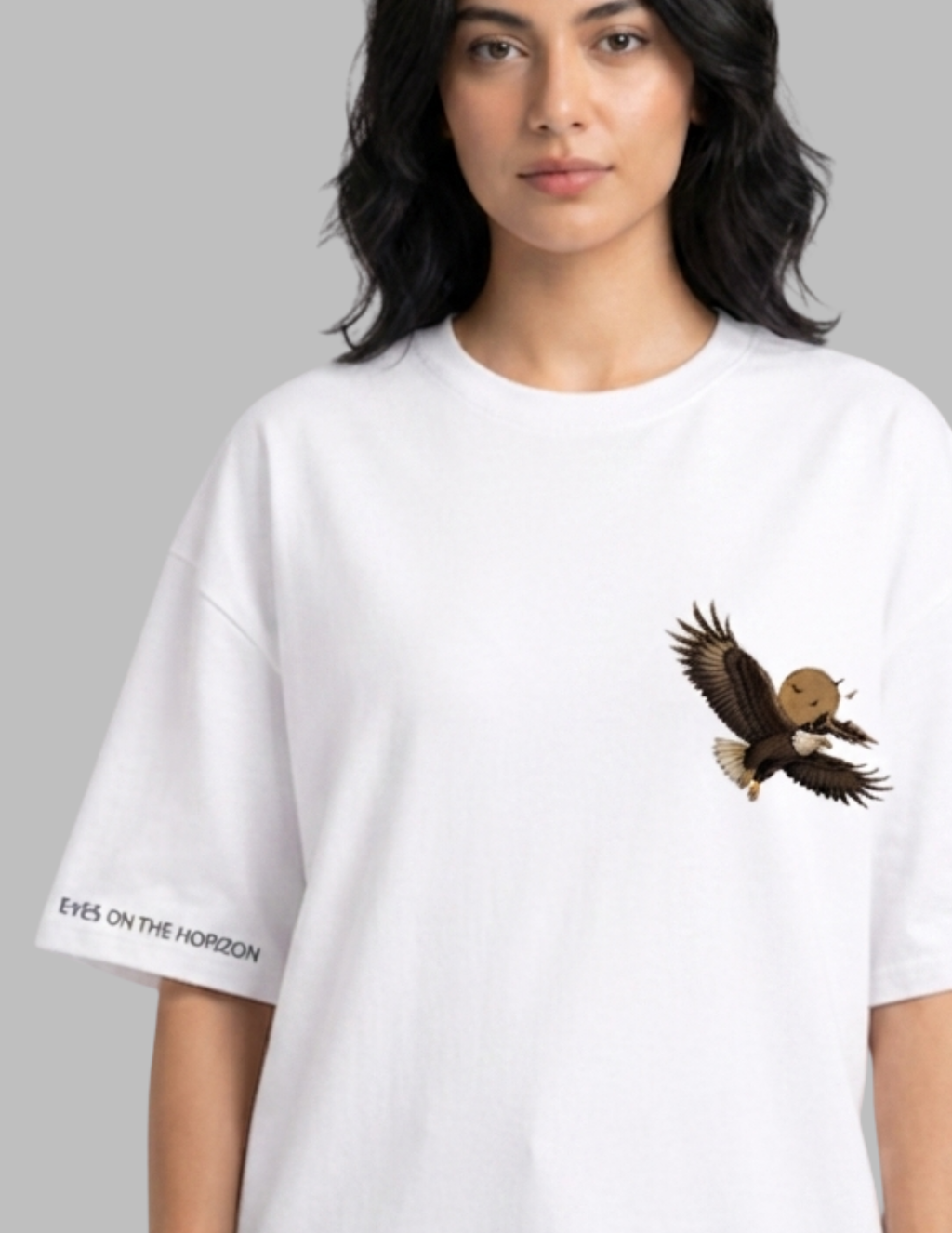 A woman wearing an oversized white t-shirt with a graphic of a flying bald eagle on the left chest and the text 'EYES ON THE HORIZON' printed on the right sleeve, set against a solid gray background.