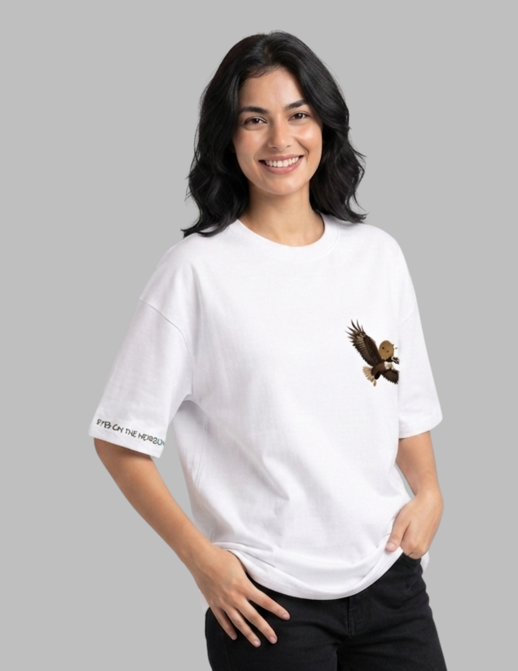 A woman wearing a white oversized t-shirt with a brown eagle graphic on the left chest and 'FISH ON THE WATER' printed on the right sleeve, set against a solid gray background.