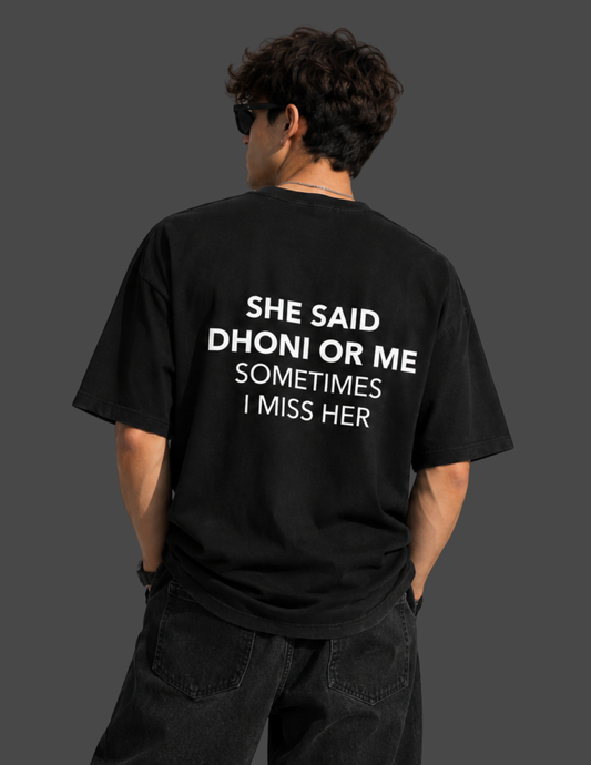 A man shown from behind wearing a black t-shirt with white text that reads 'SHE SAID DHONI OR ME SOMETIMES I MISS HER', paired with dark jeans against a dark gray background.