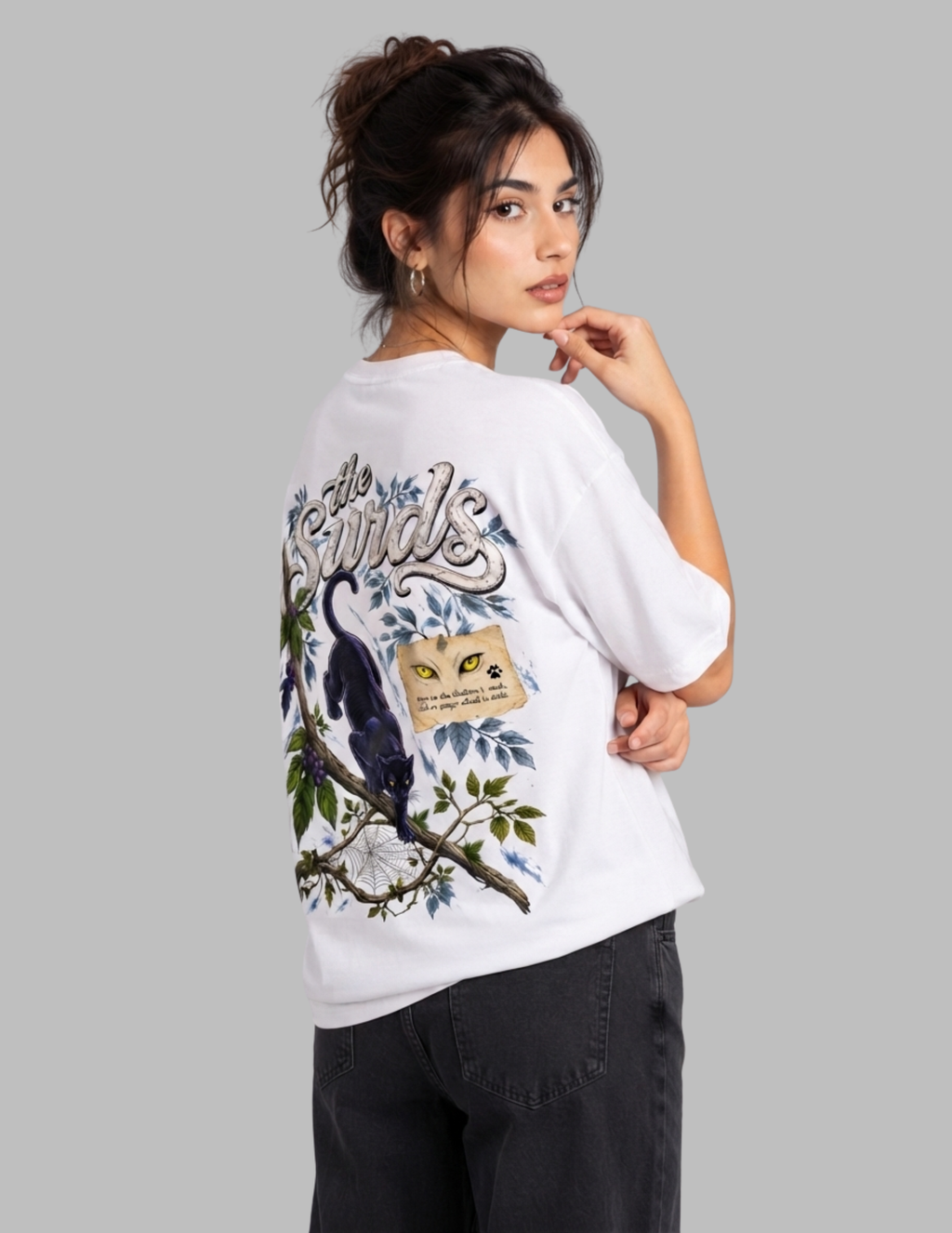 A woman modeling a white oversized T-shirt from the back, featuring a large graphic print of a bird perched on a branch with a cat's face and the word 'Finds' in stylized lettering. She is wearing dark gray jeans against a solid gray studio background.