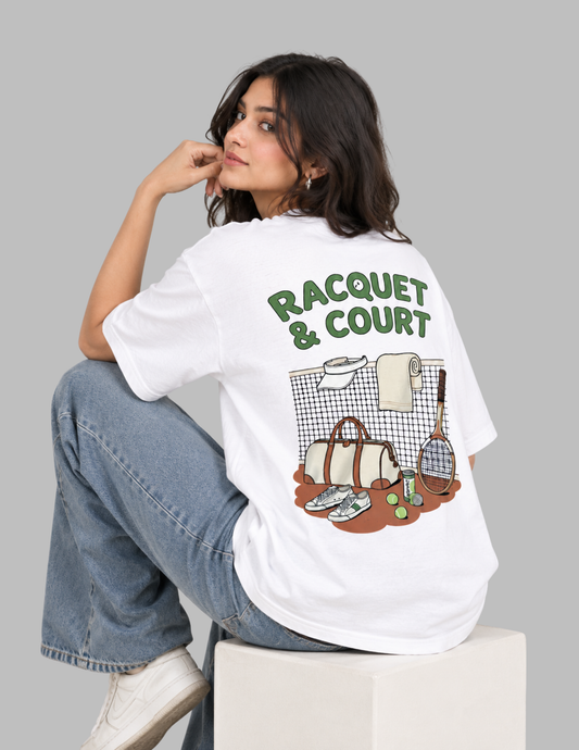 A woman sits on a white block wearing a white graphic t-shirt with a green and brown 'RACQUET & COURT' design on the back, paired with blue jeans, against a solid gray background.