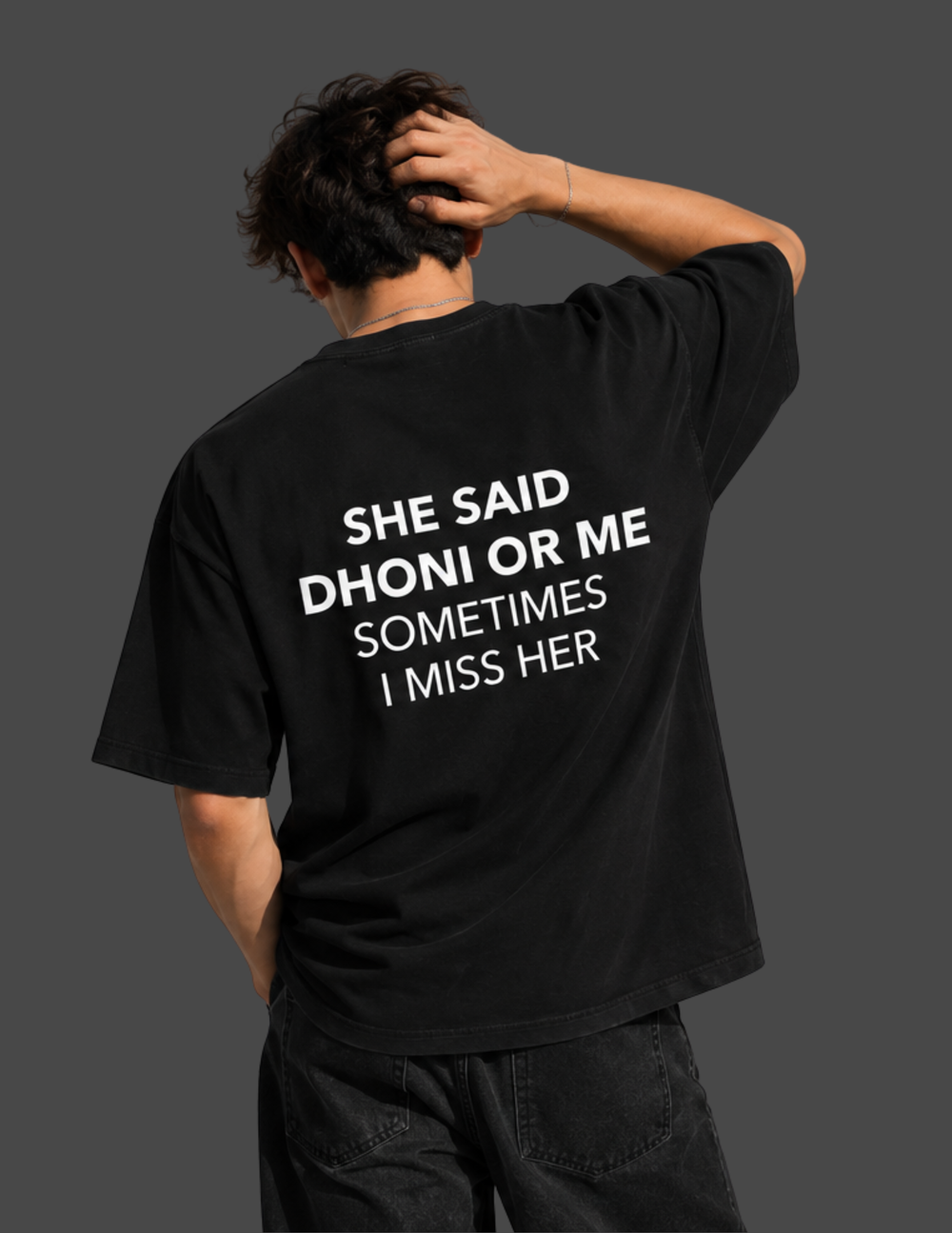 A man shown from behind wearing a black oversized t-shirt with white text that reads 'SHE SAID DHONI OR ME SOMETIMES I MISS HER', set against a dark gray background.