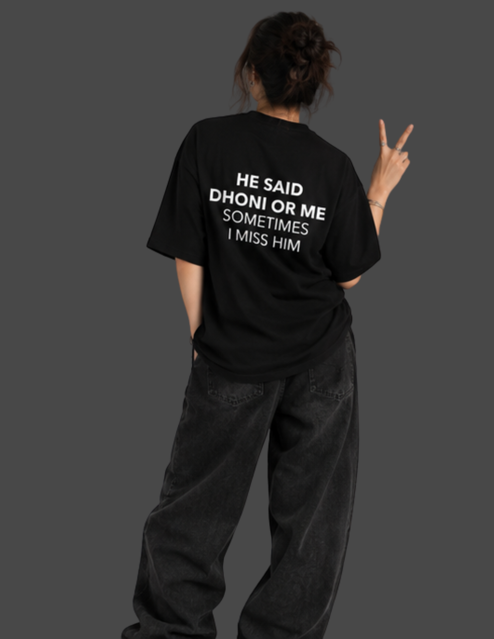 A person shown from behind wearing a black oversized T-shirt with bold white text on the back reading 'HE SAID DHONI OR ME SOMETIMES I MISS HIM', paired with loose dark jeans against a dark gray studio background.