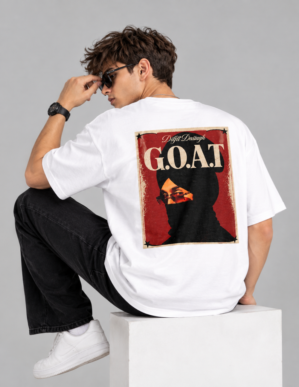 A man wearing a white oversized t-shirt with a large back graphic, sitting on a white block against a gray studio background. The graphic features a red and black portrait with the text 'Puff Daddy G.