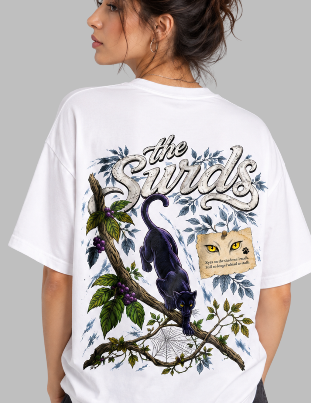 A woman wearing an oversized white graphic T-shirt, shown from the back. The shirt features a large illustration of a black cat on a tree branch with blue leaves, purple berries, a spiderweb, a paw print, and the text 'The Surds'.