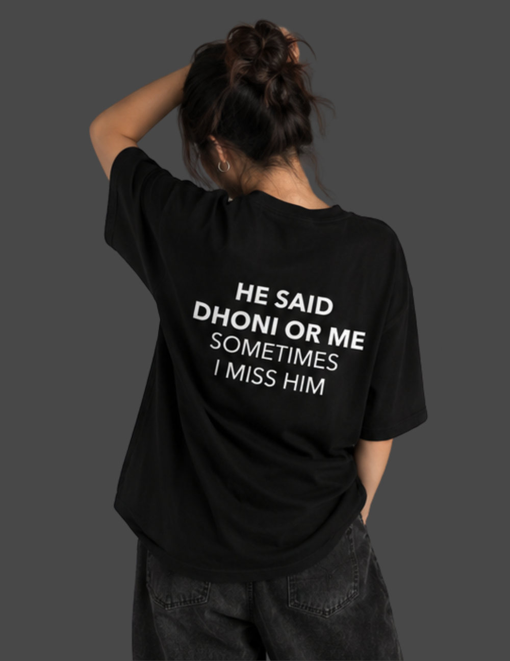 A person shown from behind wearing a black oversized T-shirt with bold white text on the back that reads, 'HE SAID DHONI OR ME SOMETIMES I MISS HIM,' paired with dark gray jeans against a dark gray studio background.