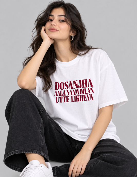 A woman wearing a white graphic t-shirt with red text that reads 'DOSANJHA AALA NAAM DILAN UTTE LIKHEYA', paired with black wide-leg jeans and white sneakers, posed against a solid gray background.