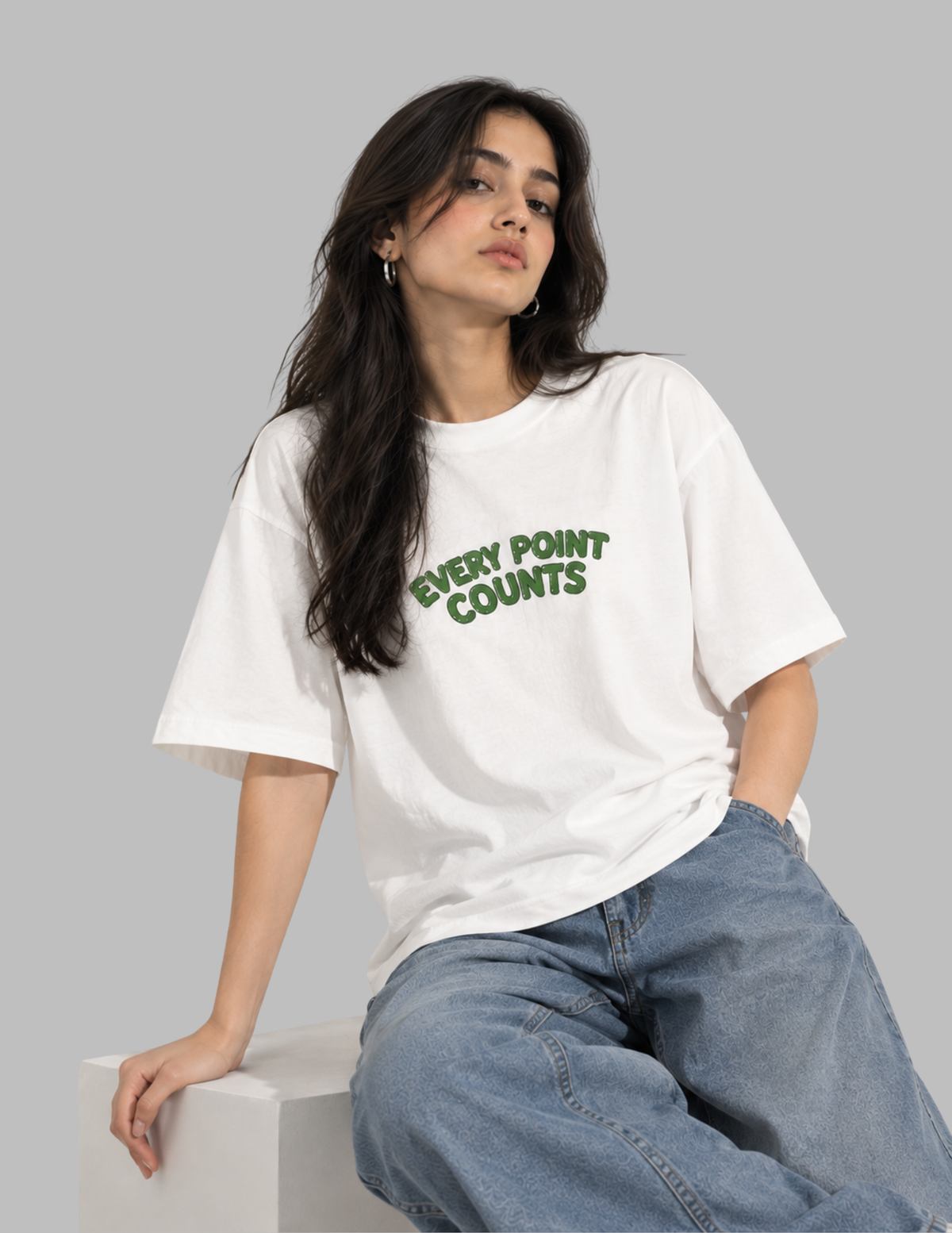 A woman wearing a white oversized graphic T-shirt with green text reading 'EVERY POINT COUNTS' and light blue jeans, seated against a plain light gray studio background.