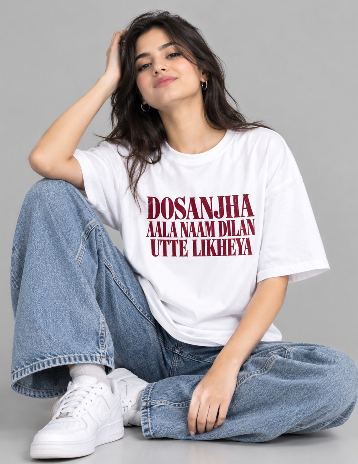 A woman sitting cross-legged on a gray studio floor wearing a white oversized graphic T-shirt with bold red text, light blue wide-leg jeans, and white sneakers.