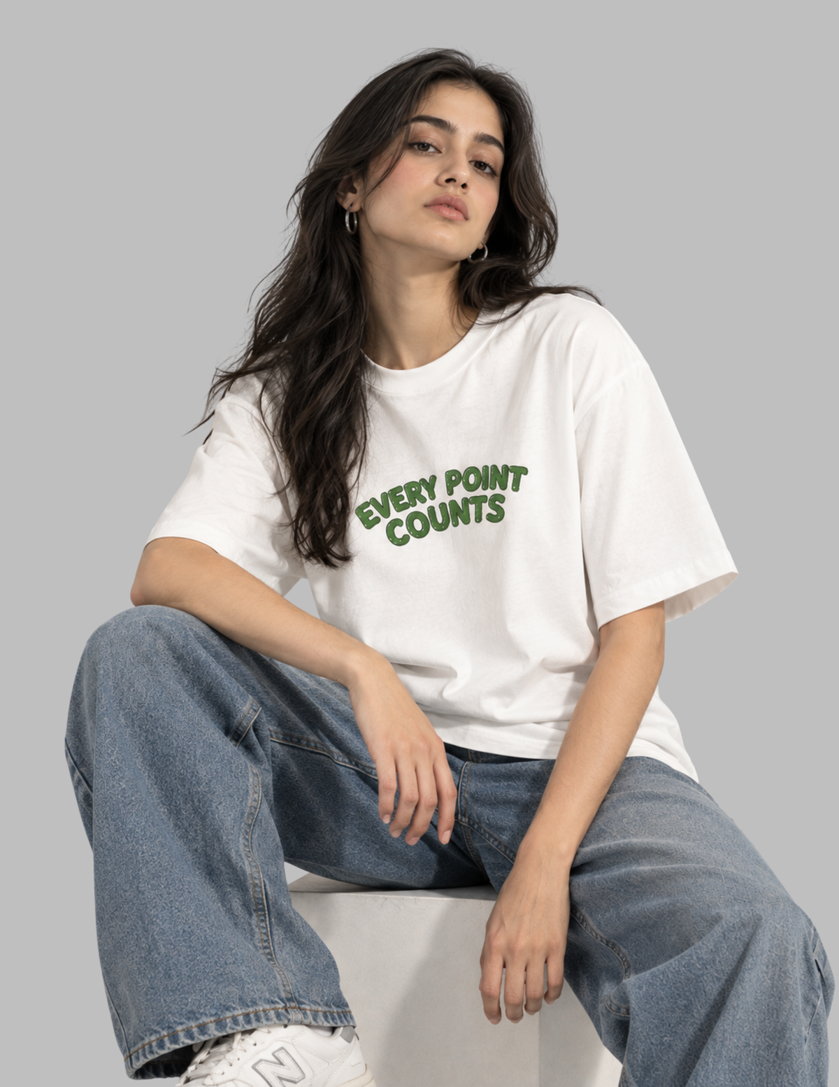 A woman sitting on a white block wearing a white graphic t-shirt with green text that reads 'EVERY POINT COUNTS' and blue jeans, against a solid gray studio background.