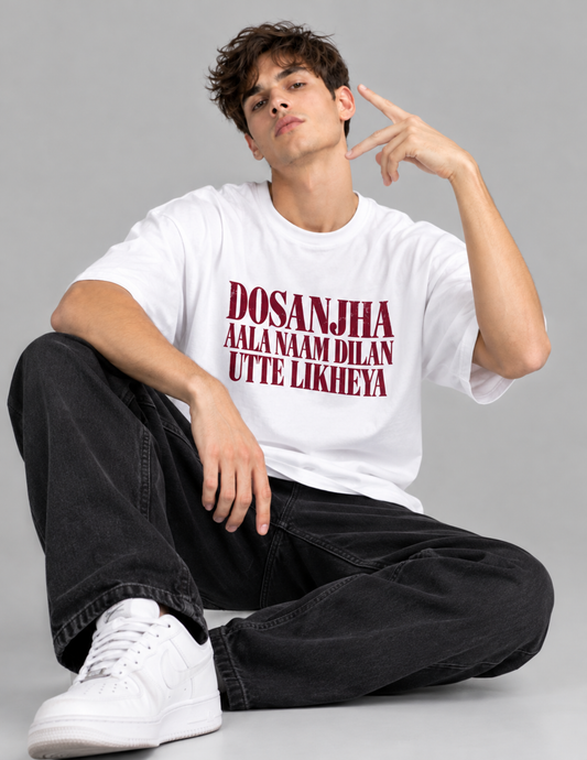 A young man sitting cross-legged on a gray studio floor, wearing an oversized white graphic T-shirt with bold red text reading 'DOSANJHA AALA NAM DILAN UTTE LIKHEYA', paired with black jeans and white sneakers.