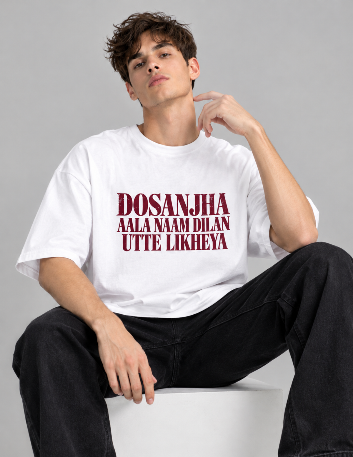 A young man sitting on a white block wearing an oversized white t-shirt with the Hindi text 'DOSANJHA AALA NAAM DILAN UTTE LIKHEYA' printed in dark red on the front, paired with black jeans, against a solid gray studio background.