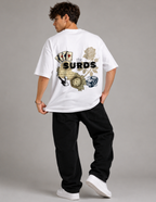 A man wearing a white t-shirt with a large graphic print on the back, paired with black pants and white sneakers, photographed from behind against a neutral gray studio background. The graphic features the text 'the SURDS' with playing cards, a rose, a clock, and dice.