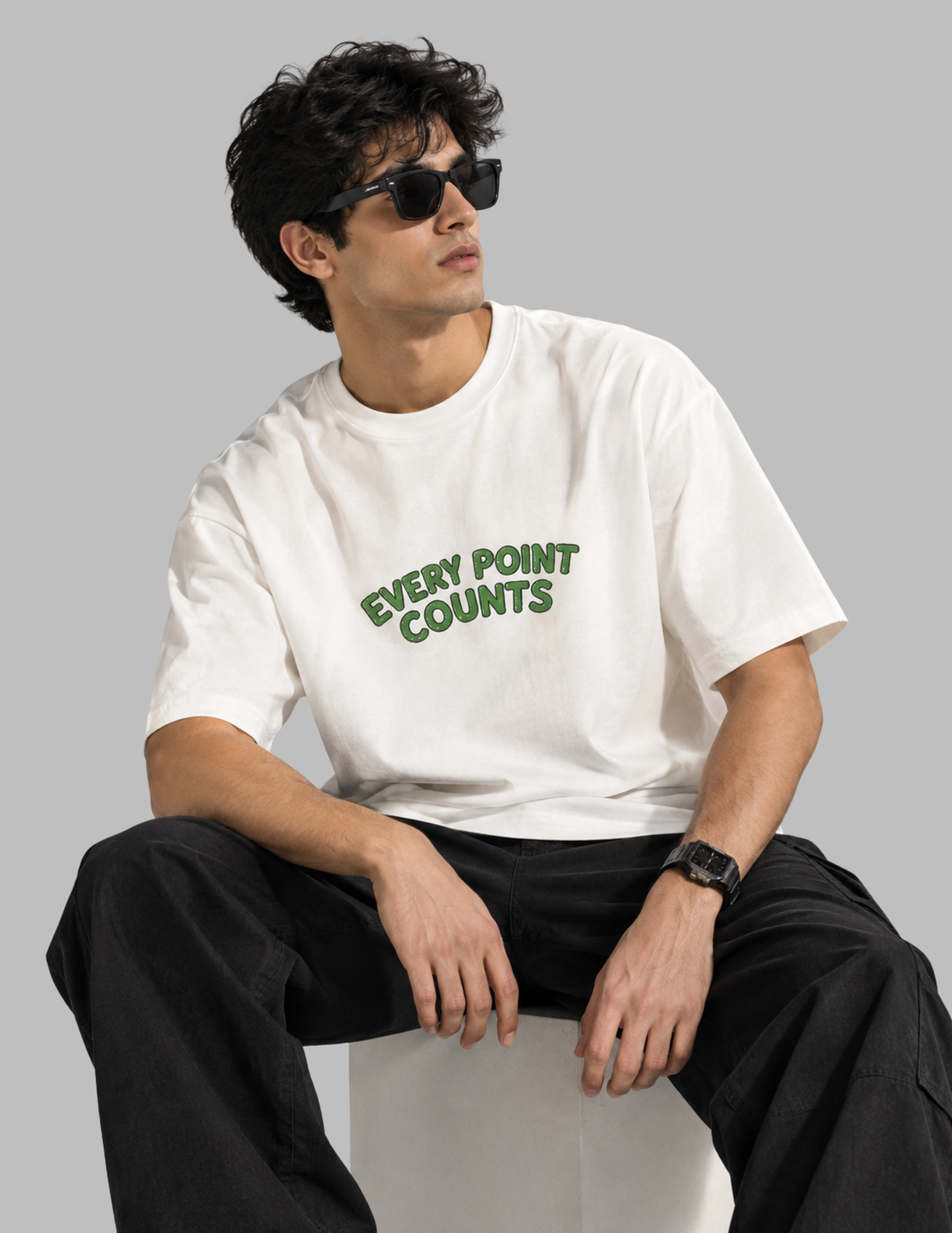A man wearing a white crewneck t-shirt with 'EVERY POINT COUNTS' printed in green across the chest, paired with black cargo pants and black sunglasses, sitting against a solid gray background.