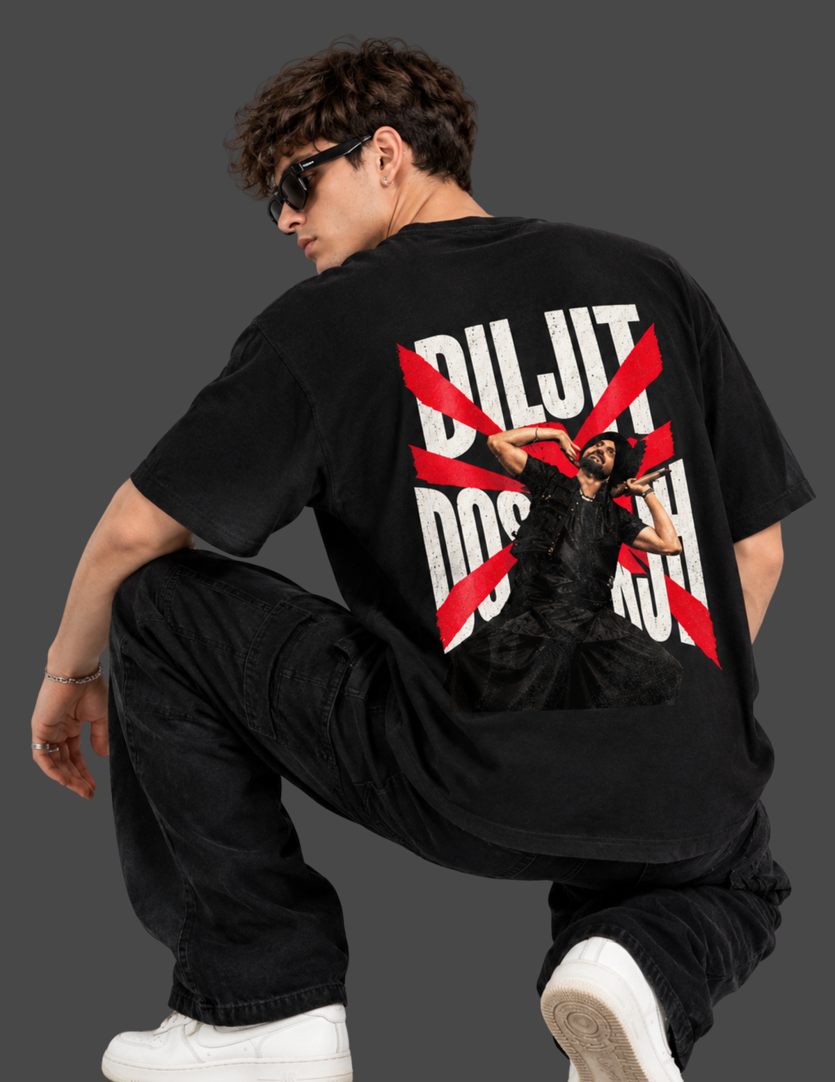 A model wearing a black oversized t-shirt with a large graphic print on the back, featuring the text 'DILJIT DOS ANCHOR' in white and red diagonal stripes, posed against a dark gray background.