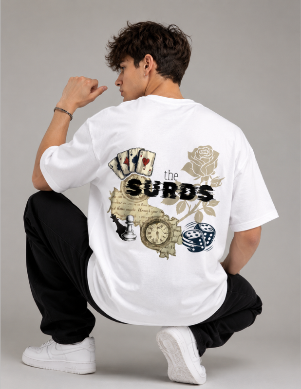 A man wearing a white oversized t-shirt with a detailed graphic on the back, crouching in a studio setting. The graphic features playing cards, a rose, a pocket watch, dice, and the text 'the SURDS'.