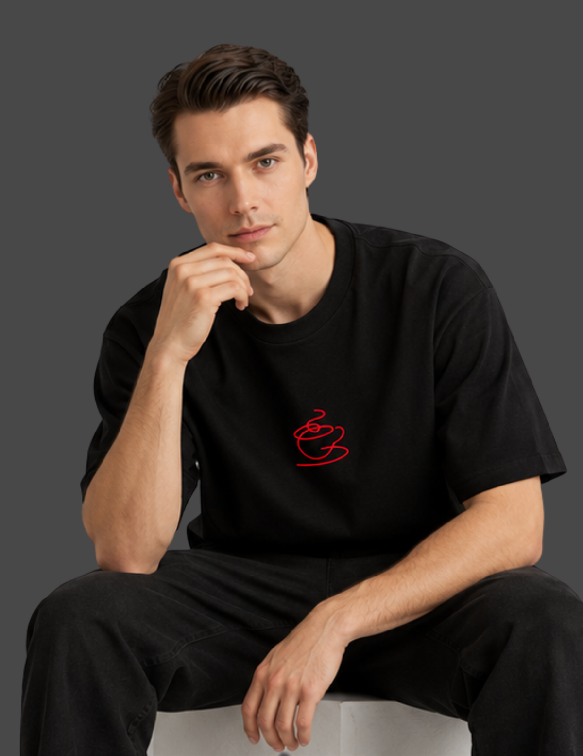 A man wearing a black short-sleeve t-shirt with a small red coffee cup graphic on the left chest, sitting against a dark gray background.