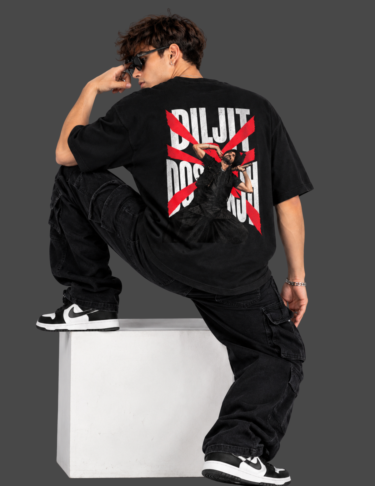 A model wearing a black oversized t-shirt with a large back graphic that reads 'DILJIT DO NOT' crossed out in red, paired with black cargo pants and black-and-white sneakers, posing on a white block against a dark gray background.