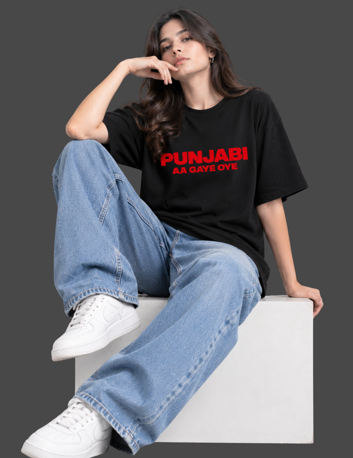 A woman sits on a white block wearing a black t-shirt with red text that reads 'PUNJABI AA GAYE OYE', paired with light blue wide-leg jeans and white sneakers, against a dark gray background.