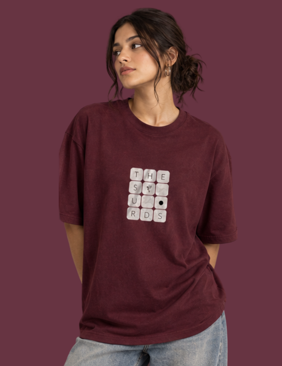 A woman wearing an oversized maroon t-shirt with a white Scrabble graphic on the front, paired with light blue jeans, set against a solid maroon background.